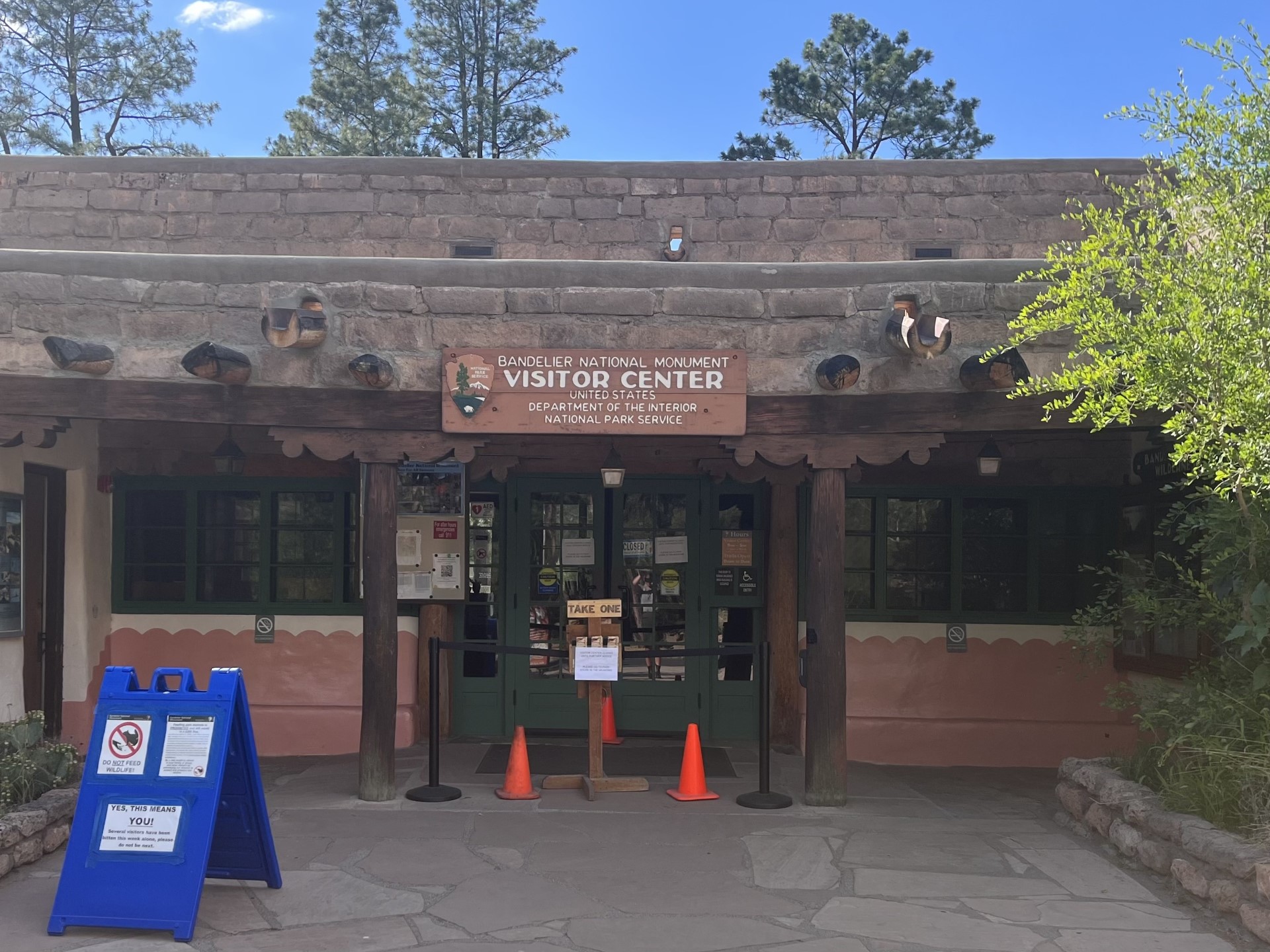 Bandelier National Monument's visitor center is temporarily closed for evaluation of environmental conditions. A stone building with green doors, which has orange cones set up in front of them. A stanchion blocks access to the building.