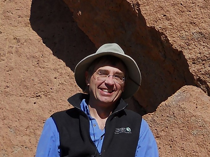 A man wearing a blue shirt and black fleece vest stands in front of a pinkish rock.