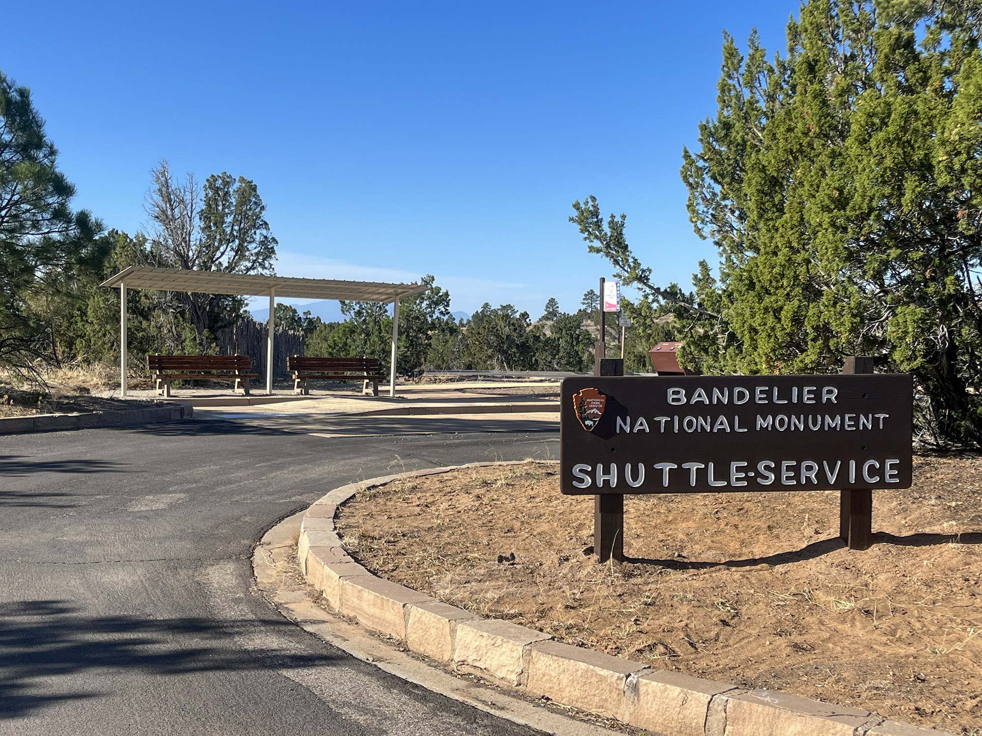 This year, the shuttle runs within the park between the Frey Trail Parking Lot and the Bandelier National Monument Visitor Center. A parking lot with a brown sign that reads "Bandelier National Monument Shuttle Service."