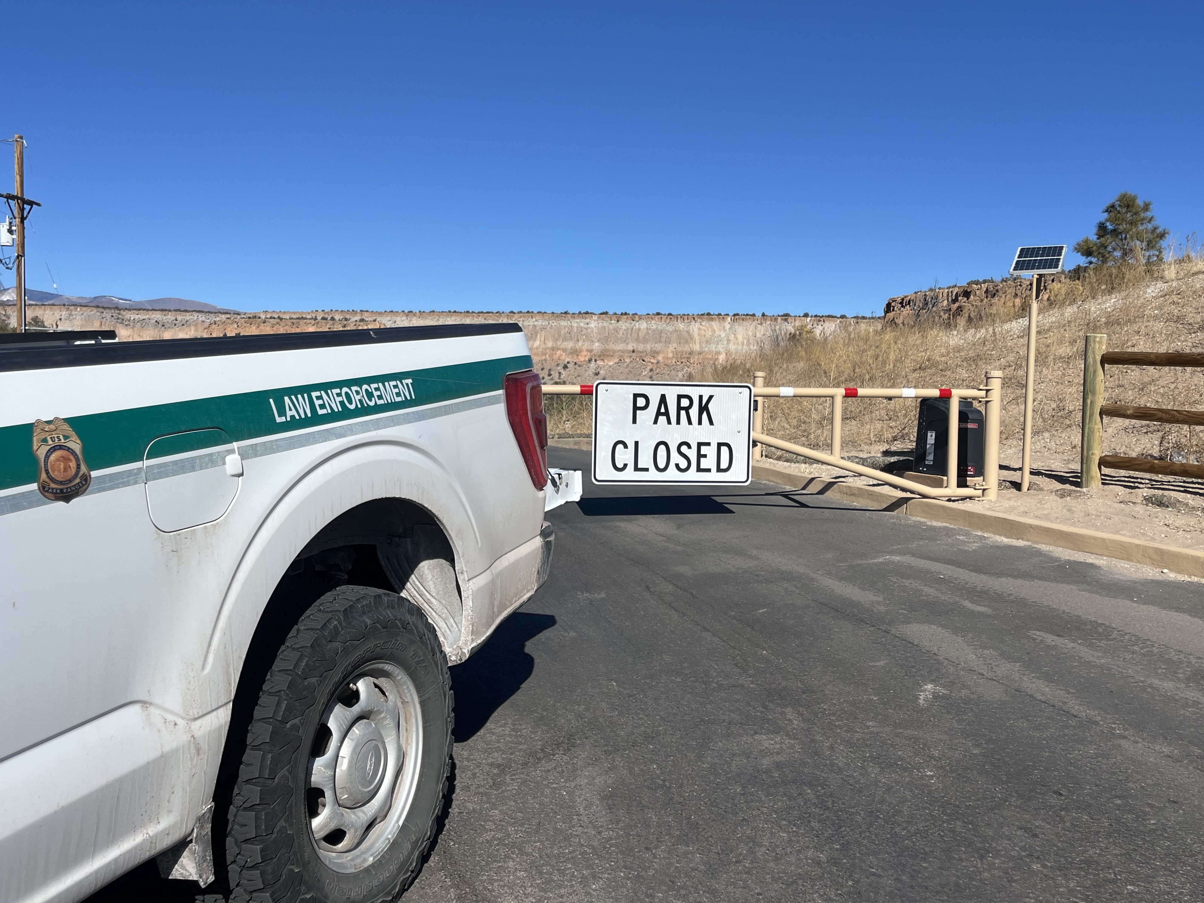 Bandelier's Tsankawi Unit is temporarily closed due to a water line break. A gate with a sign that reads "PARK CLOSED." The back end of a white truck with a green stripe is also seen.