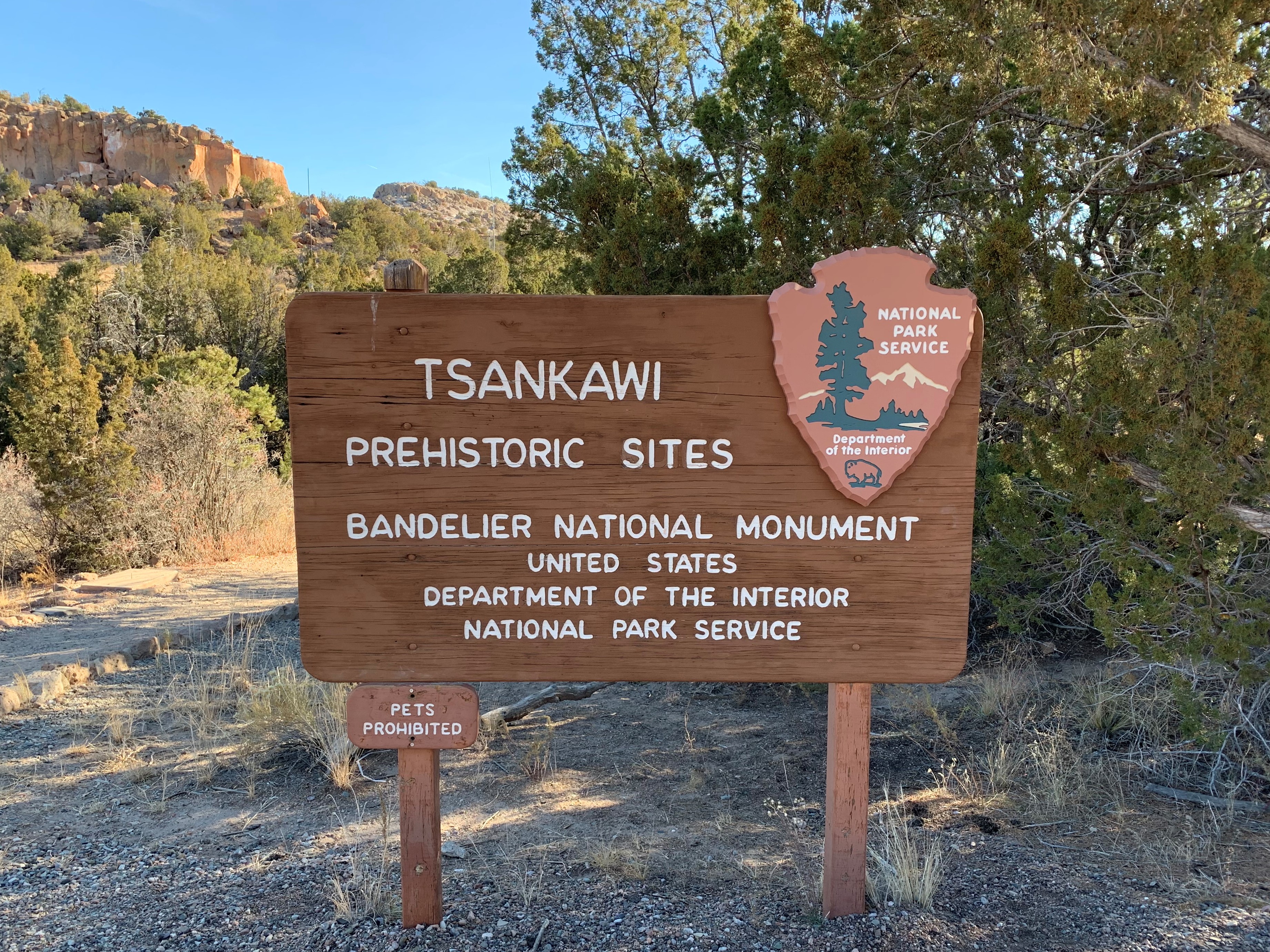 A wooden sign that says, "Tsankawi Prehistoric Sites, Bandelier National Monument, United States Department of the Interior, National Park Service." Behind it are trees with green foliage. The sky is blue.