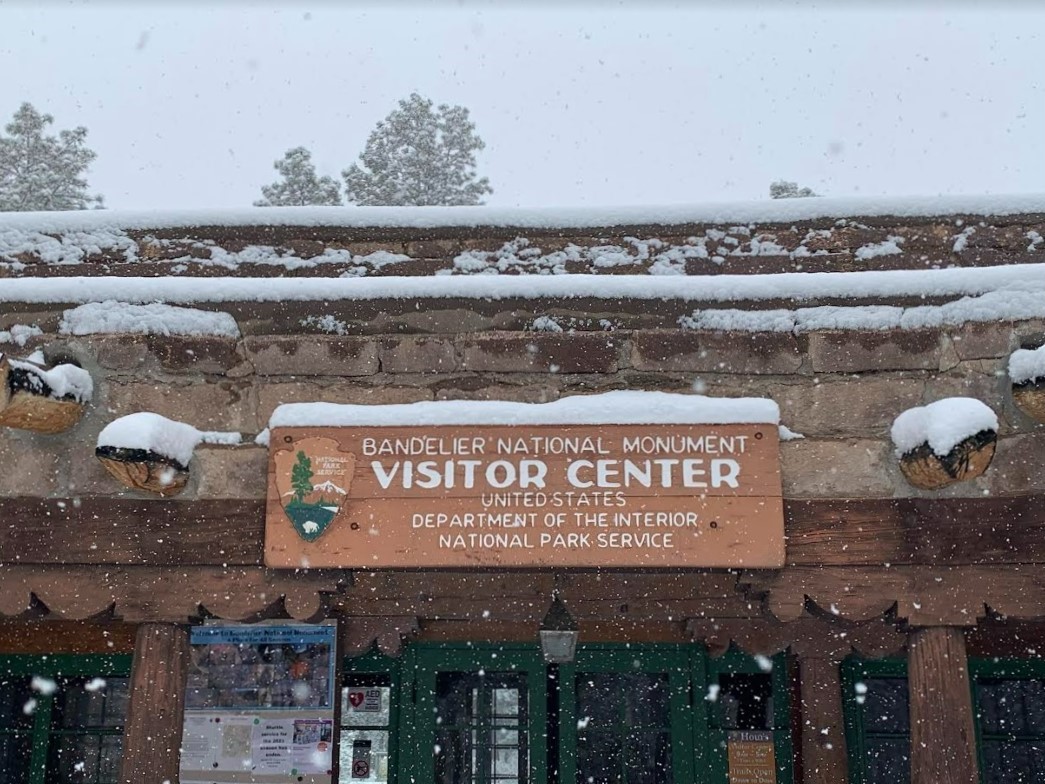 Bandelier National Monument will open at 12 noon on Sunday, February 11, 2024 to allow for park snow removal operations. A snow covered sign that reads "Bandelier National Monument Visitor Center."