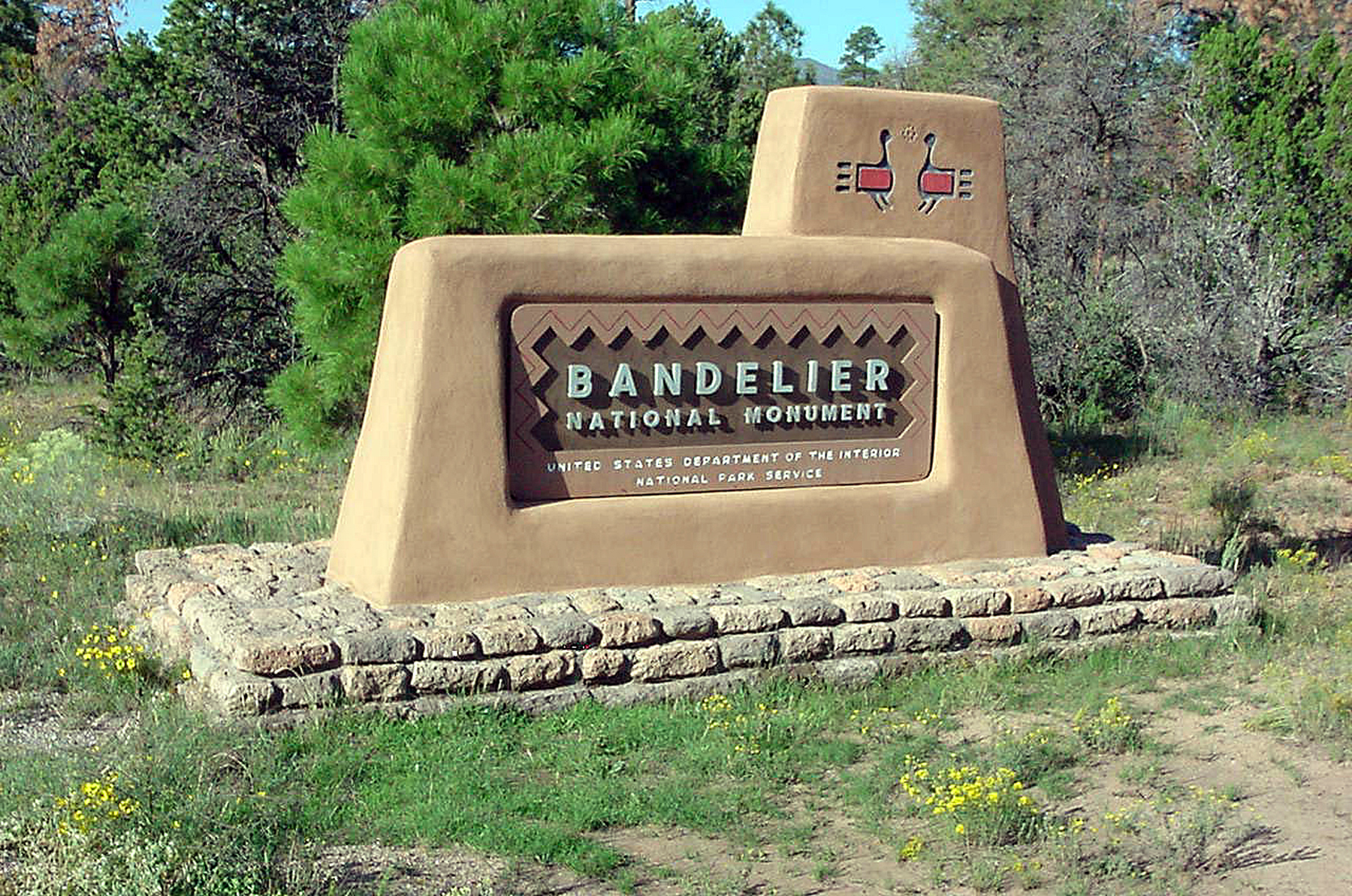 a stucco sign declares Bandelier National Monument with yellow flowers all around