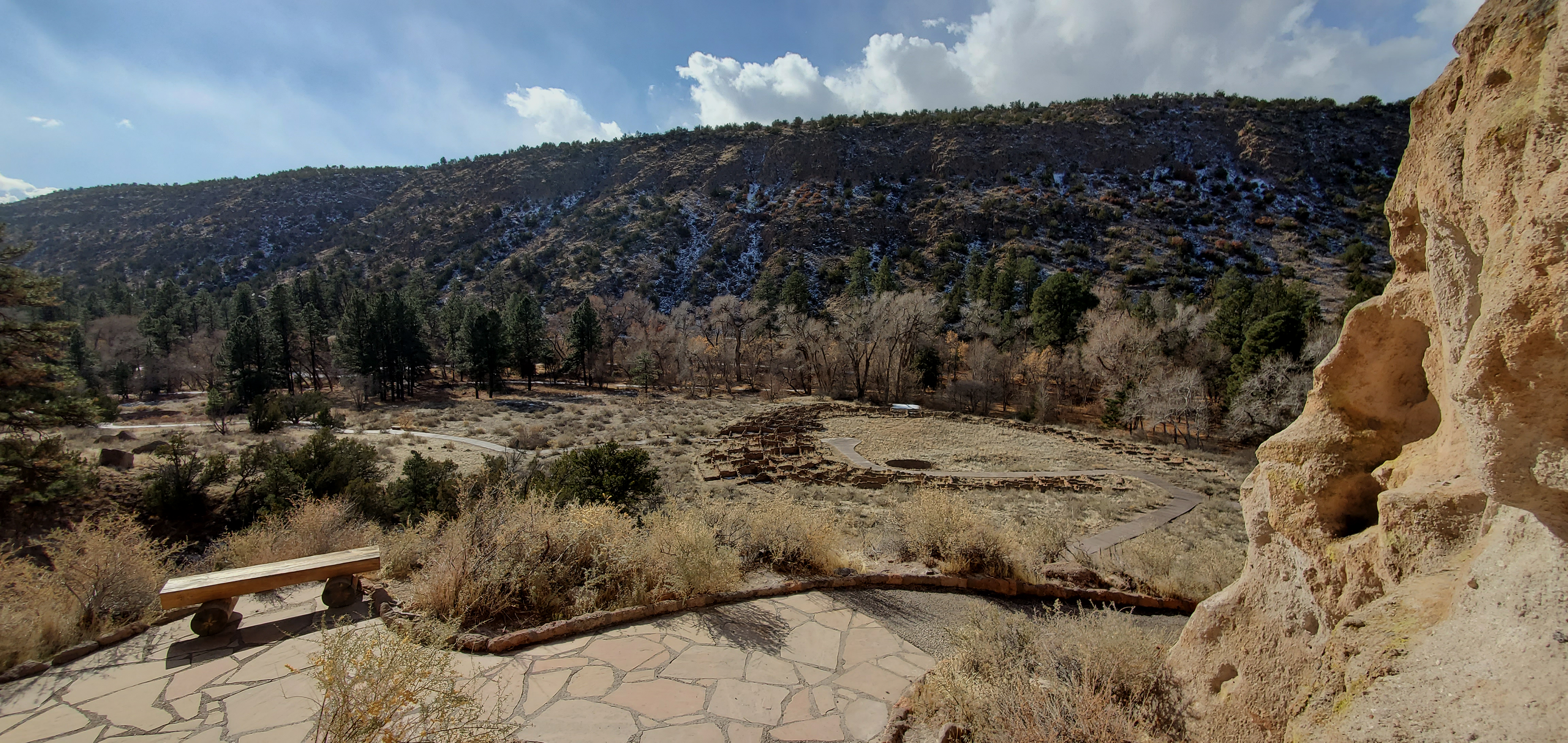 A view looking at the remains of a stone village in the bottom of a canyon with some tall green trees.  There is a bench in the foreground.