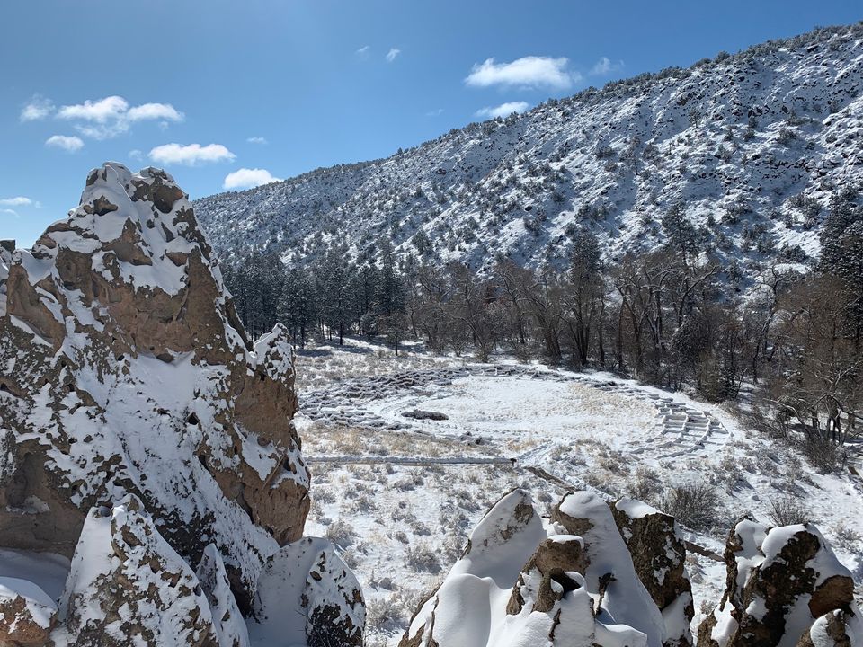tyuonyi snow White snow covers the rocks, trees, and stone foundations of a pueblo. The sky is blue with white clouds.