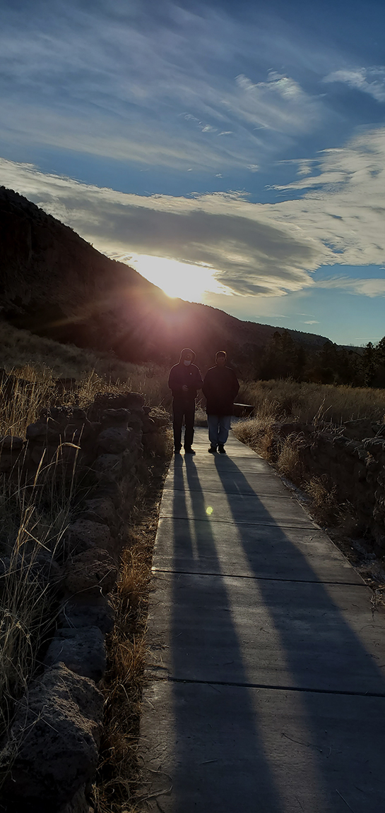 two people stand between two short stone walls with an opening, their shadows from a rising sun behind them align perfectly with the opening