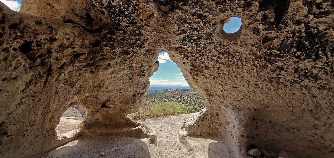 a view from inside a cavate (human made cave) with several openings that show trees and rocks outside