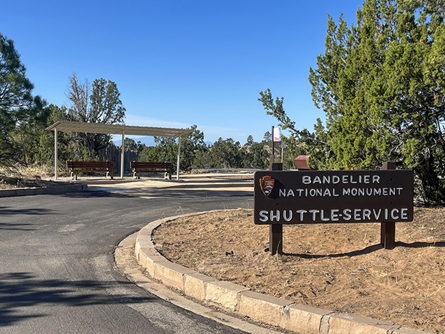 An image of a sign reading "Bandelier National Monument Shuttle Service" in a parking lot with a shelter behind it.