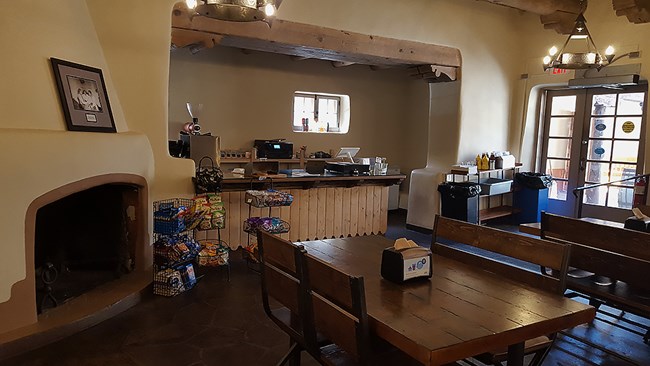 Inside of a building. There is a fireplace to the left, a counter with a cash register in the middle, and a table with chairs in the foreground. There are windows to the right.