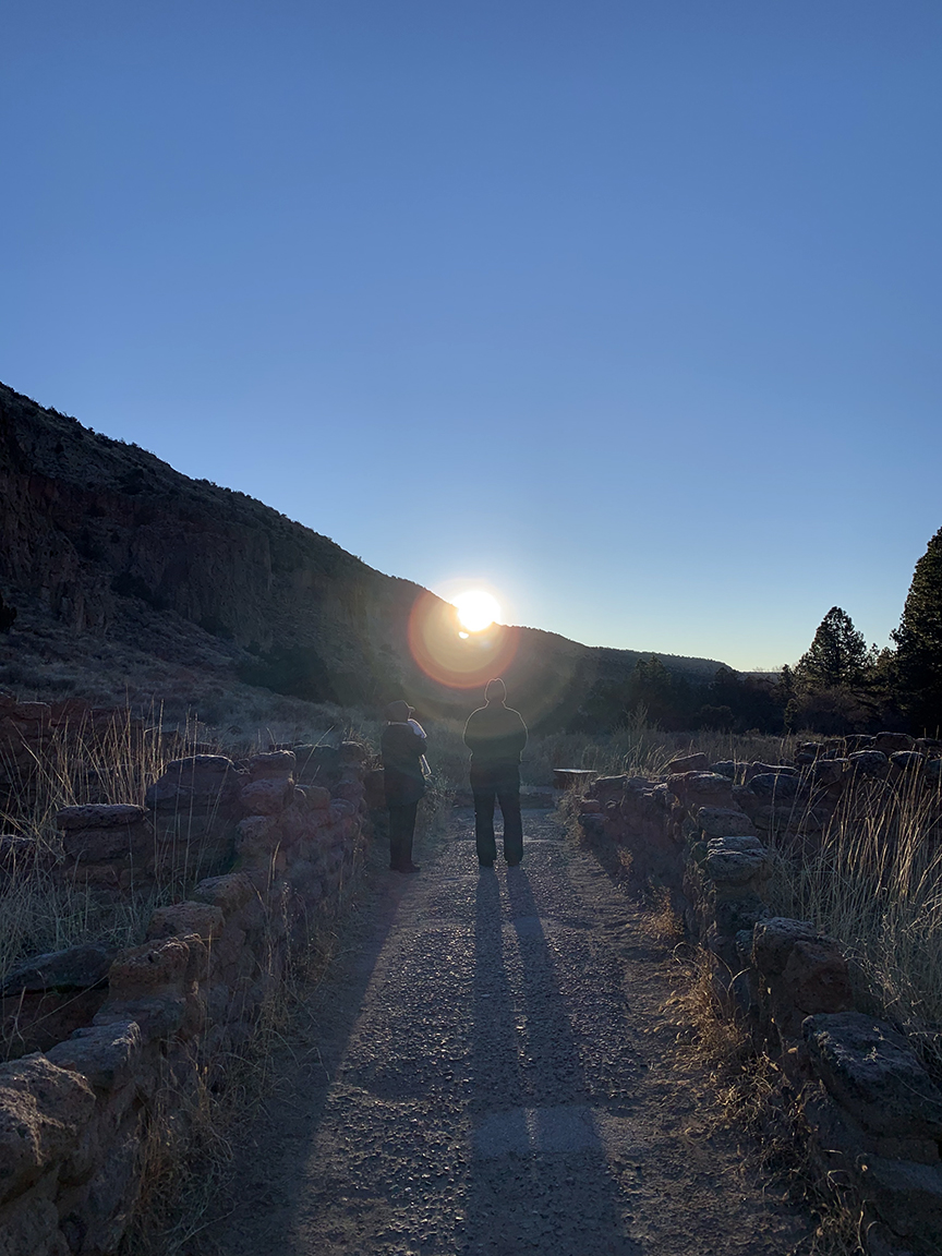 winter solstice walk 2019 Two people stand in the entrance to Tyuonyi Pueblo while the solstice sunrise directs their shadows in alignment with the pueblo entrance.