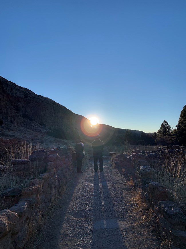 Two people stand in the entrance to Tyuonyi Pueblo while the solstice sunrise directs their shadows in alignment with the pueblo entrance.
