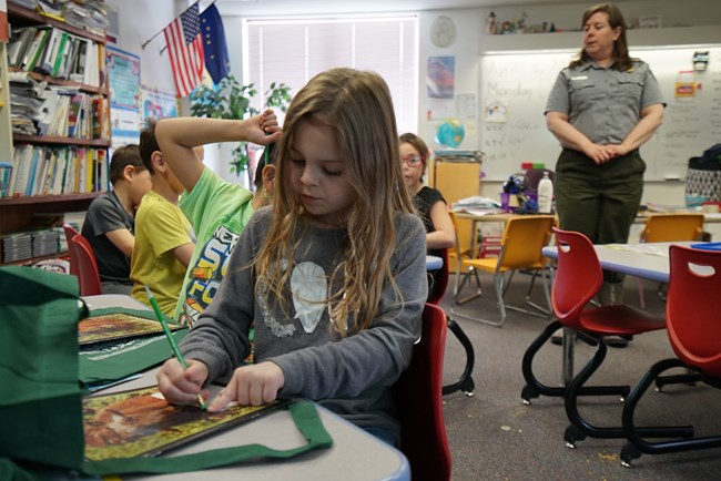 A girl holds a pencil in a classroom while a ranger looks on in the background.