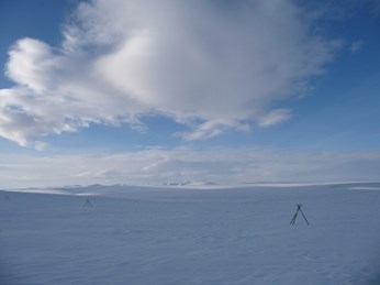 A flat, snow covered tundra with snowmobile tripod markers, under blue sky with clouds