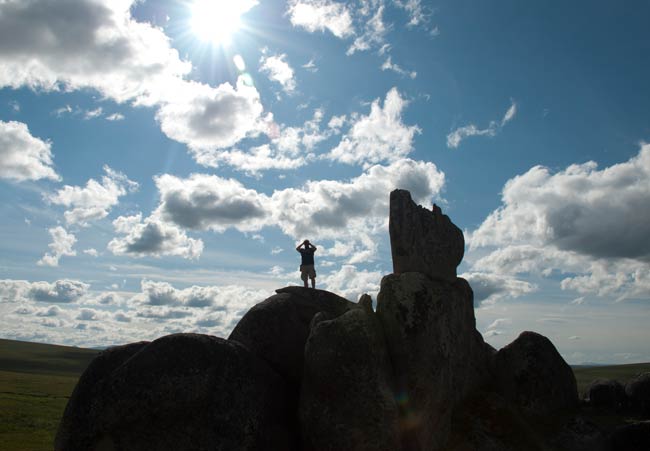 A man stands on a granite boulder under a blue sky with clouds, looking out with binoculars A man stands on a granite boulder under a blue sky with clouds, looking out with binoculars