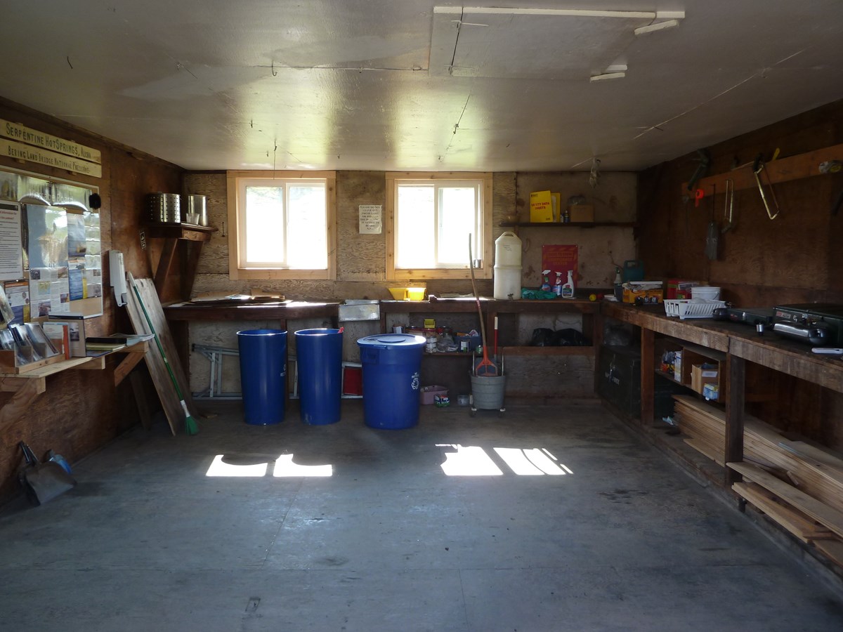 An interior room with exposed plywood walls and bare floors is lined with wooden shelving units filled with maintenance supplies and cleaning materials. Several blue trashcans, a broom and mop bucket are pushed against the wall.