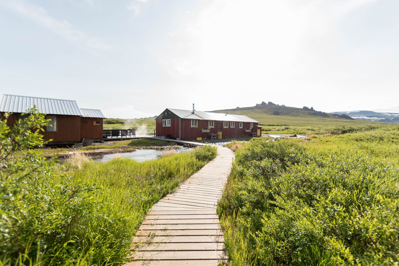 At the end of a wooden boardwalk is an enclosed bathhouse and a bunkhouse. A small creek flows beneath the boardwalk, surrounded by grasses and shrubs.