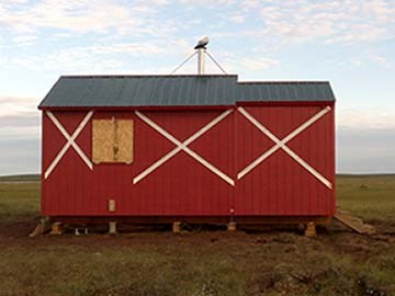 A red emergency shelter cabin with white Xs across it's length sits in the middle of a flat expanse of grassy tundra.