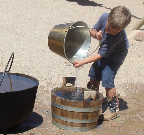 Young child fills wooden bucket with water from tin pail