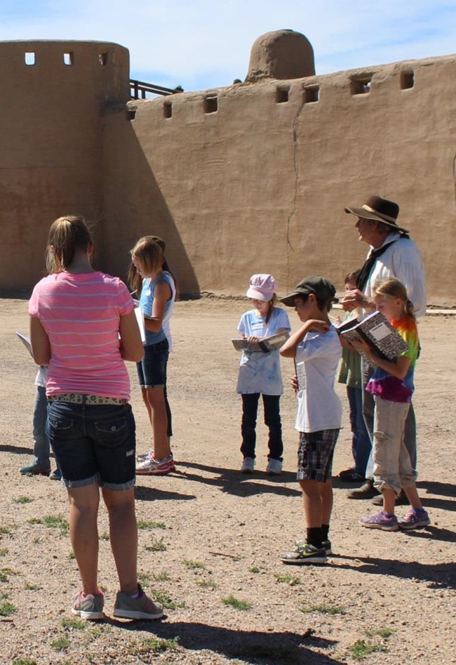 Children writing in journals with a living historian in front of Bent's Old Fort. Children writing in journals with a living historian in front of Bent's Old Fort.