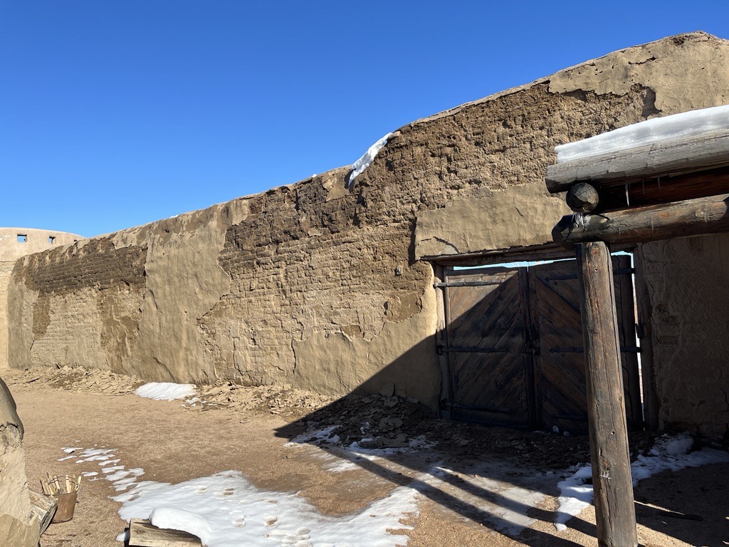 A wall of earthen brick masonry showing damage from moisture, with a pile of debris running along the length of the wall.
