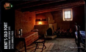 Room in adobe fort with adobe fireplace, wooden table, part of bed, window, and roof beams
