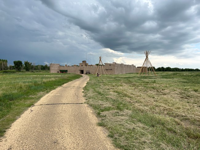 A paved path leads to an adobe fort with a tipi frame in front.
