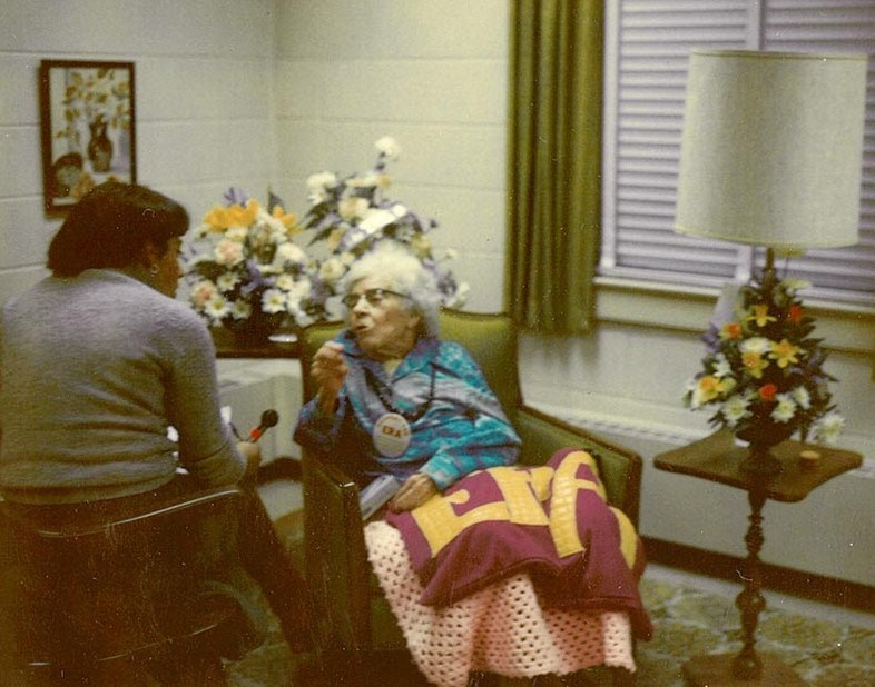 Alice Paul in wheelchair in a nursing home with ERA blanket in her lap speaking with a woman seated in a chair next to her. Bouquets of flowers are on two tables behind Paul