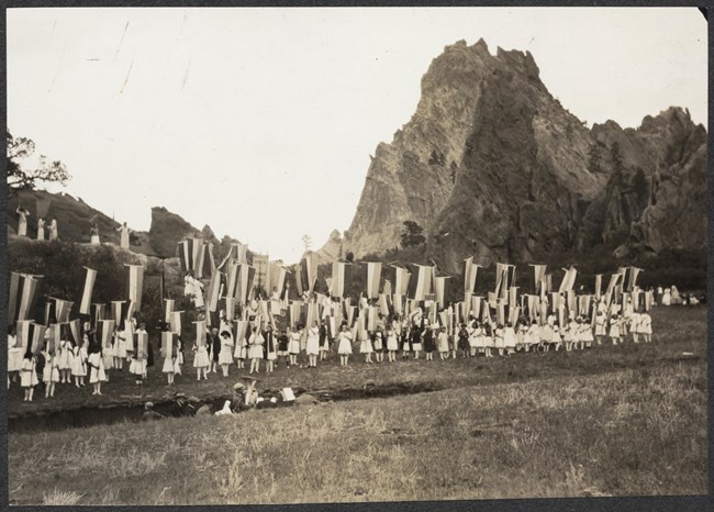 Photograph of group of girls with banners outside in front of large rock formations; sound crew and others in trench in front of them.