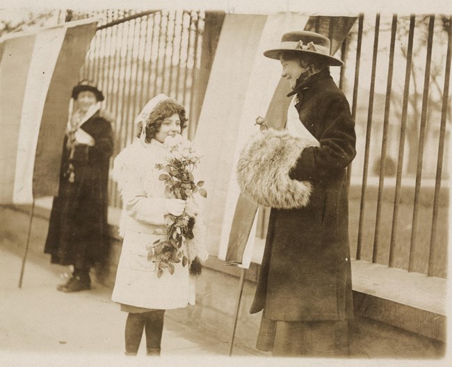 Girl giving flowers to the picketers a young girl giving a bouquet of flowers to a smiling picket with fur muff and suffrage banner as she is picketing outside the White House while another woman holding a banner looks on