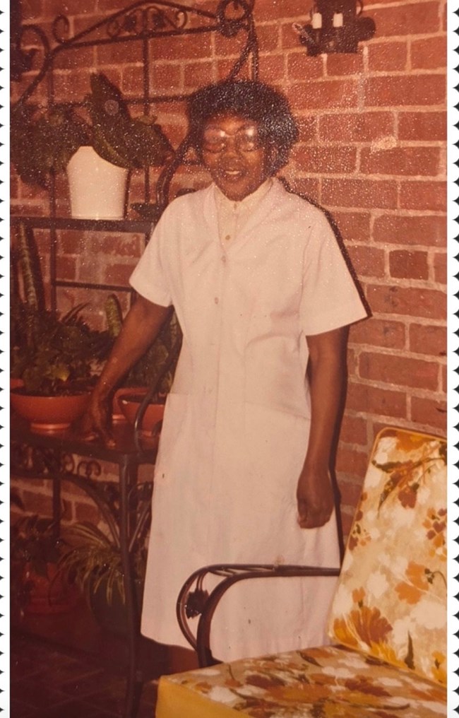 Letha Mae Glover in white dress standing next to a brick wall with slight smile and right hand resting on metal shelving unit with plants. A cushioned patio chair with yellow floral print is partially visible in foreground