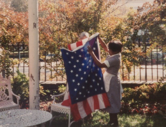 Letha wearing a maid uniform putting up an American flag on an outdoor flagpole
