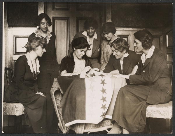 Photograph of six National Woman's Party members gathered around and watching Alice Paul stitching ratification flag.