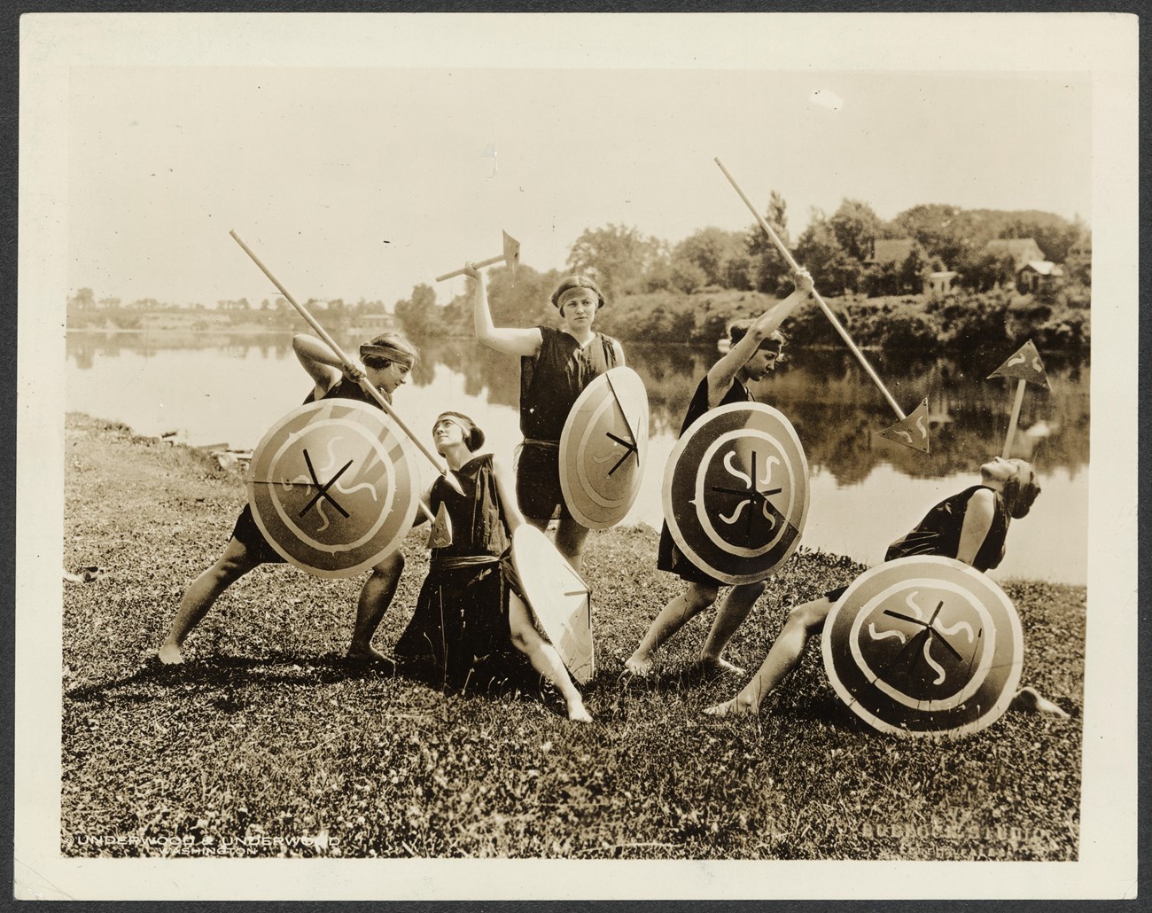 Photograph of five young women, in costume and holding shields and spears, posing in front of a lake.
