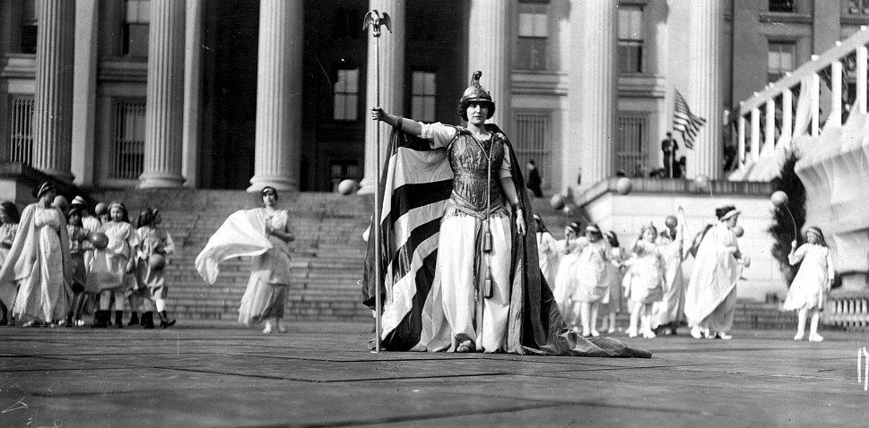 Woman wearing costume of "Columbia" with other suffrage pageant participants standing in background in front of the Treasury Building