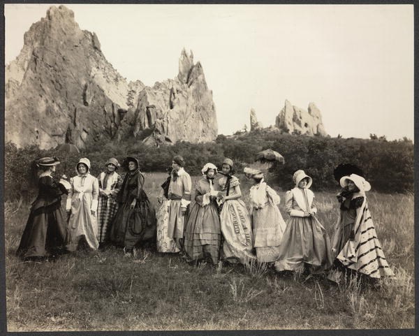 Photograph of group of women in nineteenth-century costume standing outside, large rock formation in background.