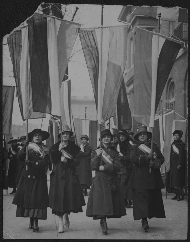 Working Women's Picket Four women wearing coats, hats, and suffrage sashes carrying tri-colored banners