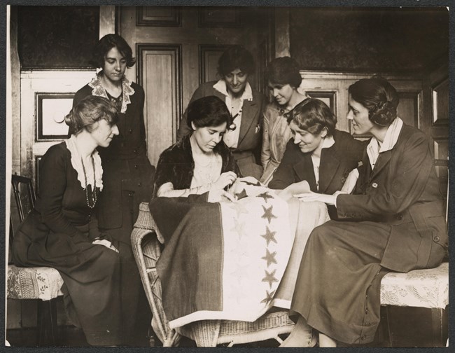 Alice Paul sits at a table sewing a star on a ratification banner while other women of the NWP look on