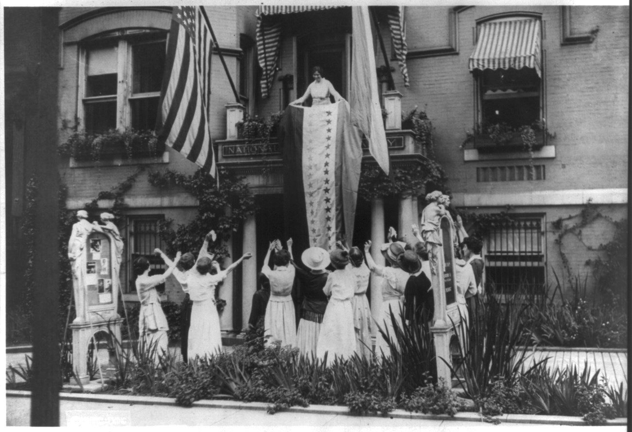 Alice Paul hangs a large tricolored banner from the balcony of the National Woman's Party headquarters. A dozen suffragists cheer on the ground below.