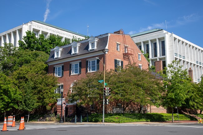 A four-story brick building at the corner of an intersection surrounced by trees and a tall white building behind it.