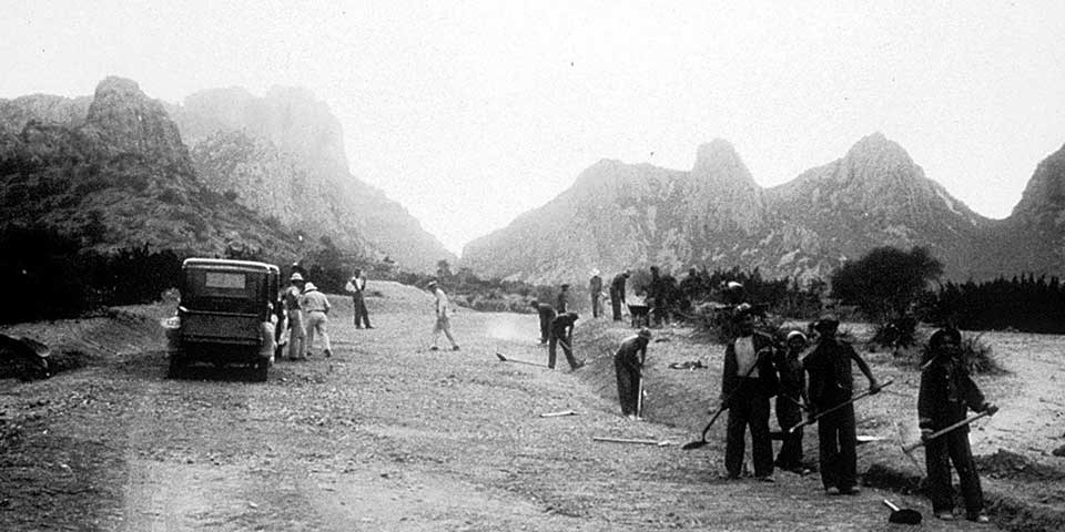 Workers with shovels build a mountain road, as shown in a 1930s photo.