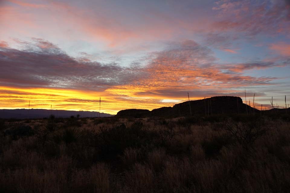 The rising sun colors the clouds gold and pink over a desert rock hill.