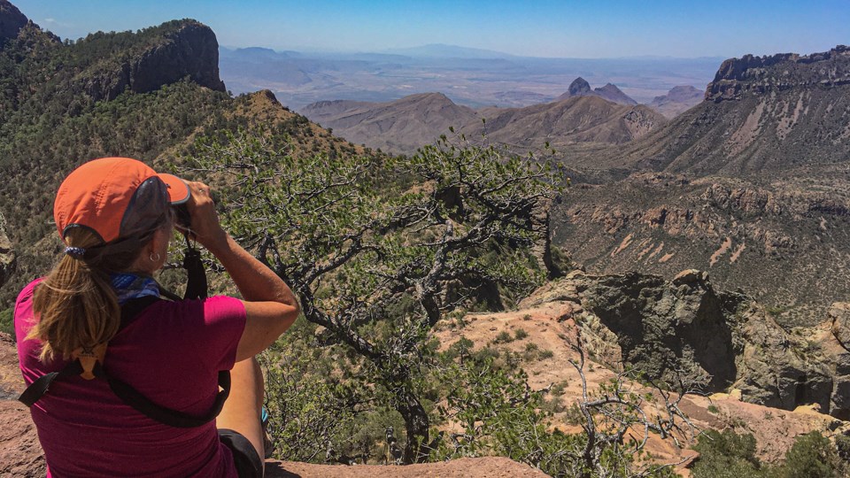 A woman sits on a rock, looking out over a vast view with her binoculars