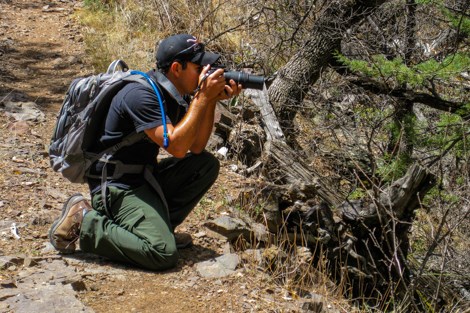 A man crouches on a trail, with his camera raised to his eye.