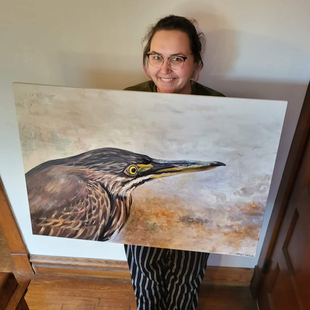 FB_IMG_1726241055258 Lindsey Winslow holds one of her finished paintings in her studio. The canvas painting depicts a bird on a foggy background with brown and white feathers, gold under beak and black top beak and golden eyes.
