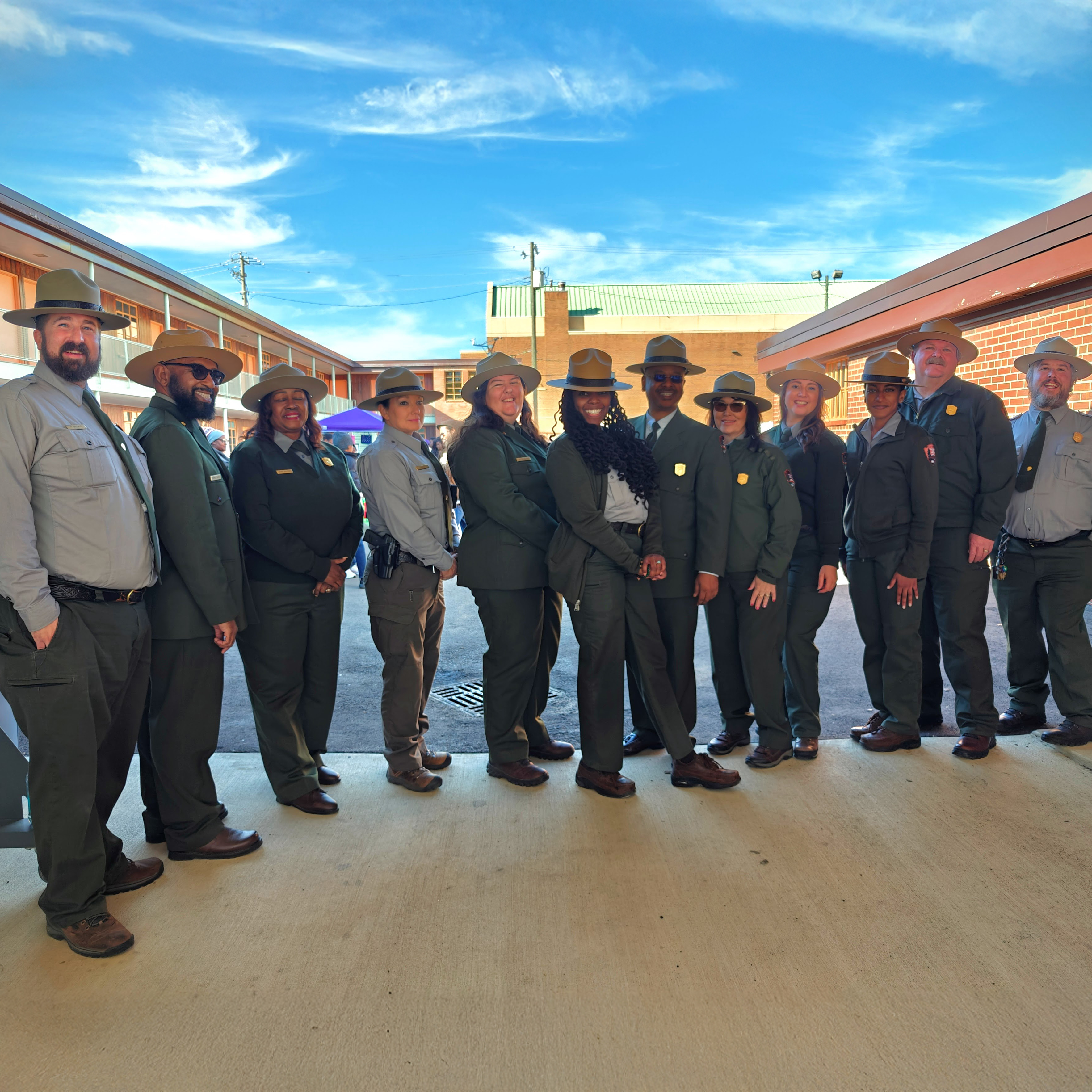 A group of park rangers stand in a line facing the camera. A brick building is seen on both sides of them.