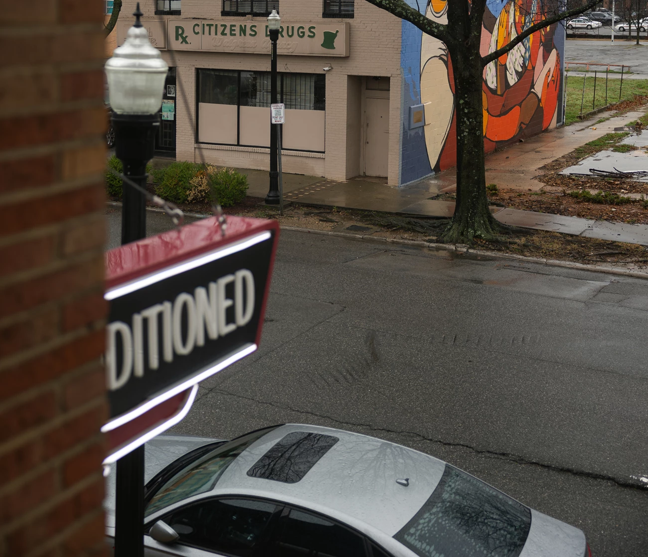 View of 5th Ave. with Citizens Drugs A street with pointed street lamps. Across the street is a building labeled Citizens Drugs with a mural painted on the side. Part of a car and a sign can be seen at the bottom of the image.