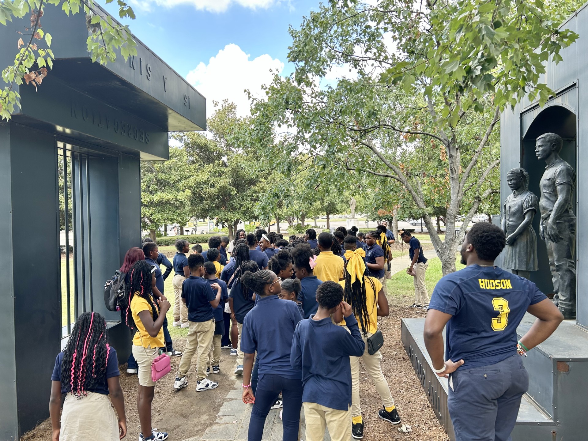 A group of students listen to a ranger giving a program in a park. A statue of metal bars is on the left side and a metal statue of children is on the right side.