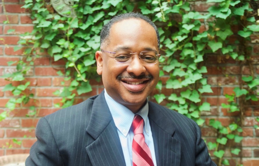 Rolando A well dressed man in a suit and tie smiles at the camera in front of a brick wall covered in green ivy.