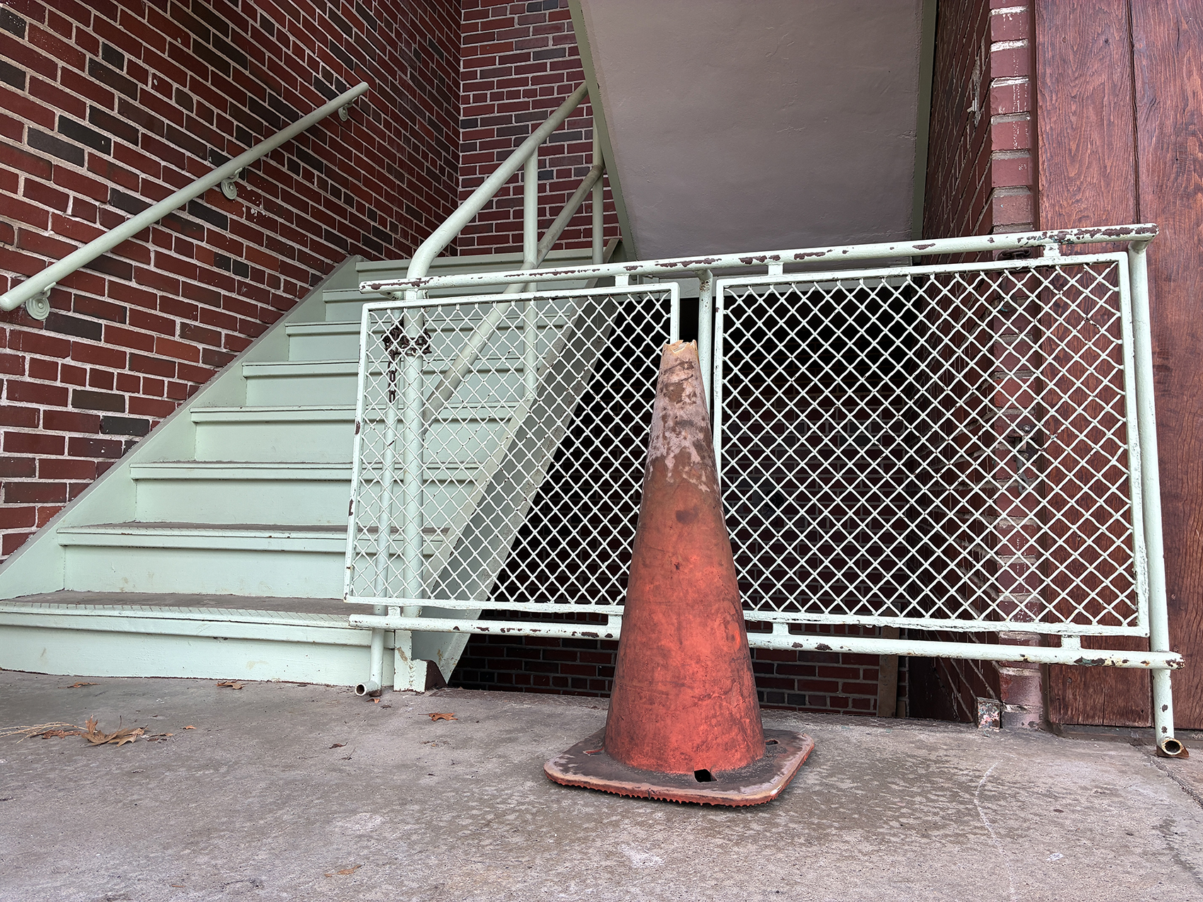 A plastic safety cone sits in front of a metal barricade of a staircase going downstairs. Stairs going up a brick building are located left of the barricade.