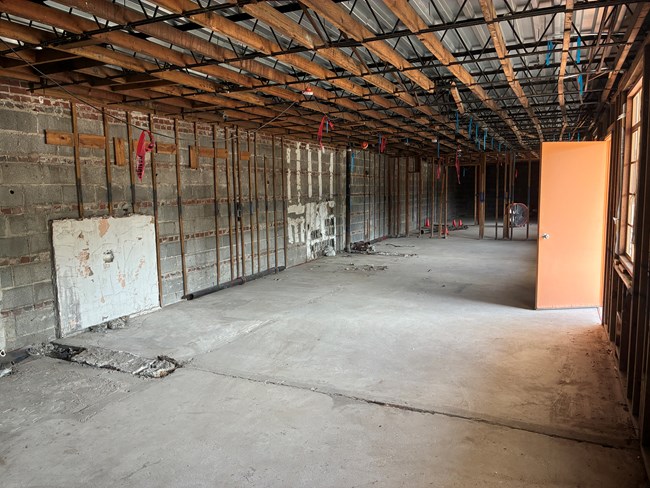 An empty upstairs room ready for the next round of renovations. Brick walls with some wood remaining is on the left side of the image. Bare concrete floor and ceiling are visible. In the background a remnant wall framing remains standing.